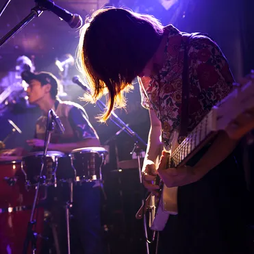 A guitarist looks at their hands with their hair covering their face as they play guitar on stage next to a microphone with a drummer in the background and moody purple lighting