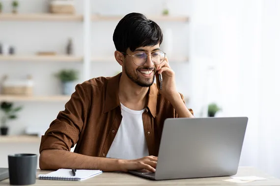 Cheerful student sitting at desk with laptop having phone conversation, typing on computer keyboard