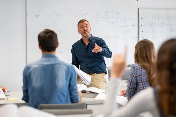 An academic at the front of a classroom in front of a whiteboard addresses a student with their hand raised sat in the foreground near two of their classmates