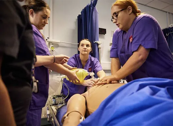 Three student nurses performing CPR on a mannequin in a simulated hospital environment.