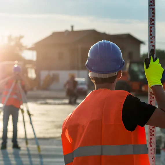 Two surveyors in blue hard hats and orange high visibility jackets use land surveying and measuring equipment on a construction site outside