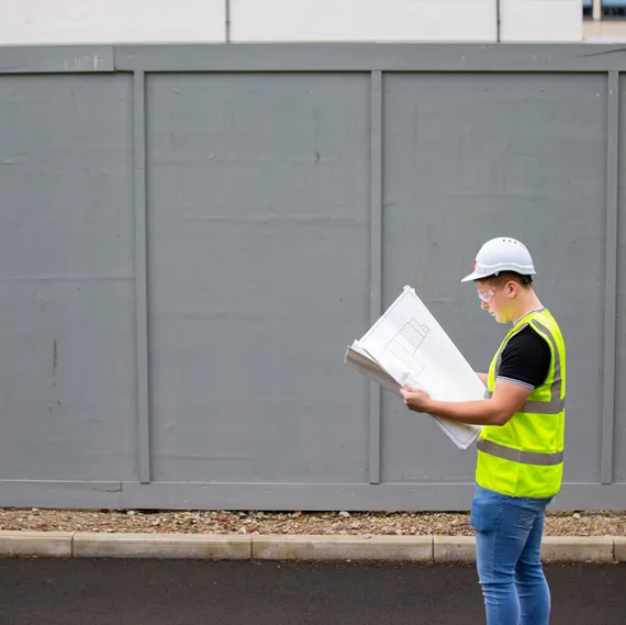 A student wearing a white USW hard had and a yellow high visibility vest examines blueprints on large white paper at a building site in Treforest