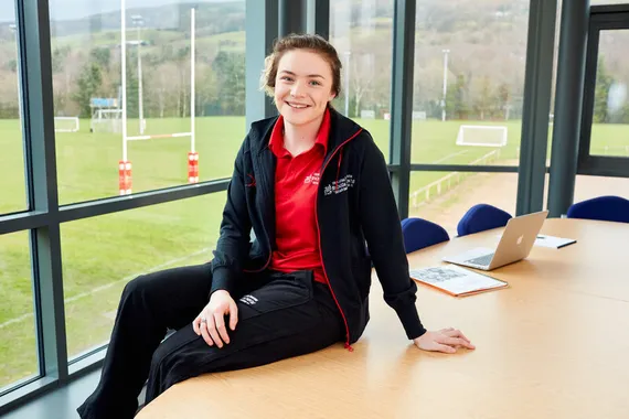Sport student, Ffion Denley, is sat on a table smiling at the camera. In the background, you can see USW's outdoor sports pitches.