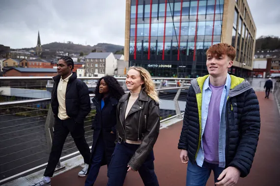 Four students walk together in a row on a footbridge over the river Taff in Pontyrpidd.