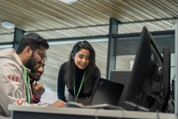 Three students of the global majority smile while chatting around a computer at Newport campus