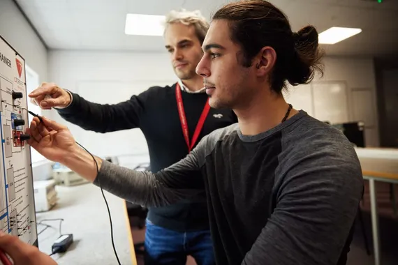 Automotive Engineering student Alexios Papazoglou conducts an experiment on an electrical board attached to a wall while seated at a desk and being supervised by a lecturer