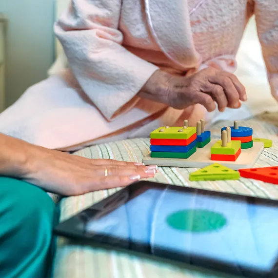 A health assistant in green scrubs uses wooden toys and a tablet with an elderly dementia patient while sat on a bed