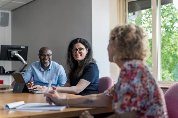 Three members of staff, who are part of Ethnic Minority Research Advisory Group (EMRAG), sit around a table smiling and chatting with notepads and pens