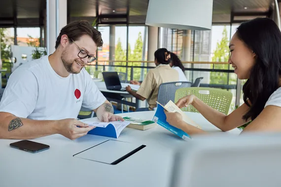 Two students chatting over books while making notes at a table in Newport campus