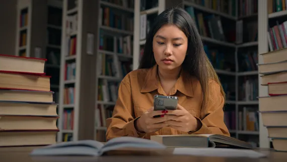 A student looks serious while looking at their phone in a library with a pile of books next to them, an open book in front of them and bookshelves behind them