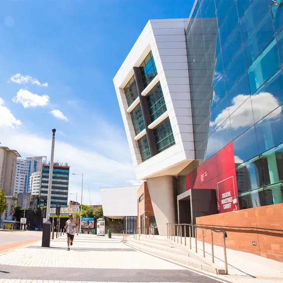 A right-side view of the front entrance to Cardiff Campus with a blue sky and student accommodation buildings to the left and a person running on the pavement outside