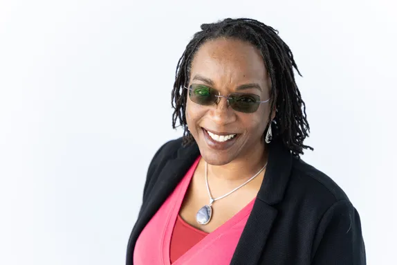 Sociology professor Dr Roiyah Saltus smiles at the camera while wearing sunglasses in front of a white backdrop