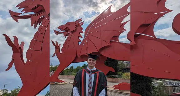 Graduate James McFarlane dressed in a suit, graduation robes and a graduation hat proudly stood in front of the big red dragon outside Newport's ICC
