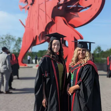 Graduates Robyn Kelly and Amy Bartlett pose in front of the red dragon outside Newport's ICC wearing graduation robes and caps