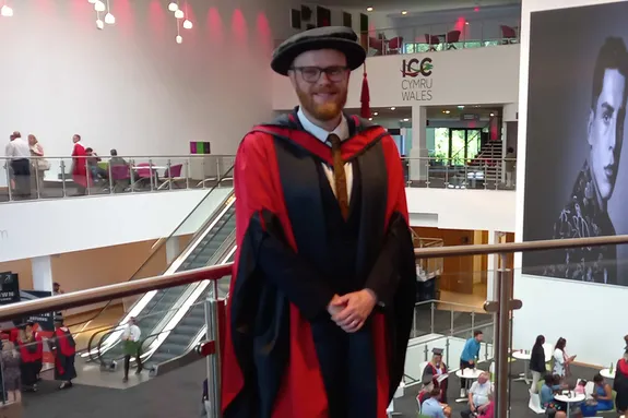 Huw Stephens is stood smiling wearing a graduation cap and gown.