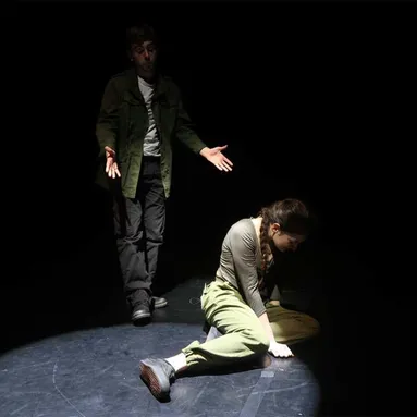A sad-looking woman sits in a spotlight on the stage floor staring at the ground while a man stands behind her with outstretched hands talking
