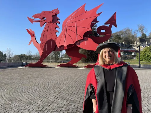 A woman smiling wearing a black and red graduation gown and hat. She is stood in front of a large red metal dragon with bright blue sky