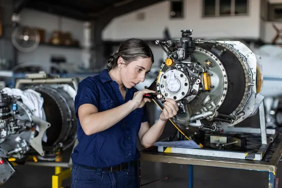 A person in a blue shirt uses a tool to work on a large mechanical component in an indoor industrial setting.