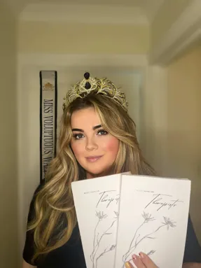 A woman smiling holding up two books in her hand with a white front cover. She has a gold crown on her head.