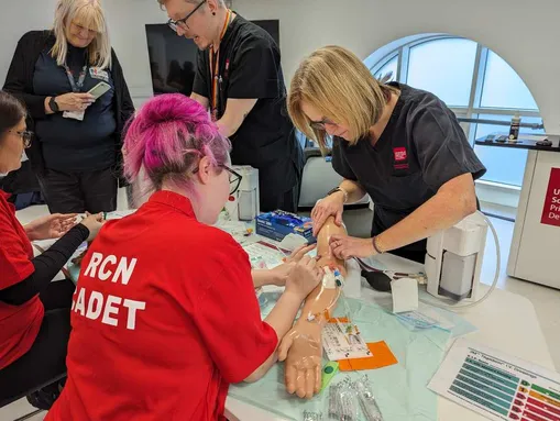 Medical trainees practice IV insertion on a mannequin arm under the guidance of an instructor in a medical training setting
