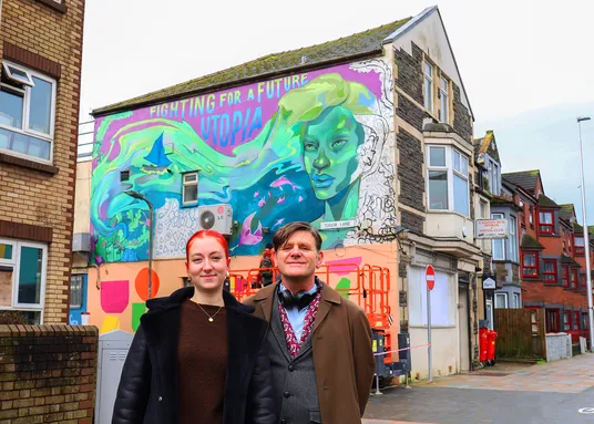 Two people are stood on the street. A house behind them has a large colourful mural featuring a woman's face. It reads 'fighting for a future utopia'