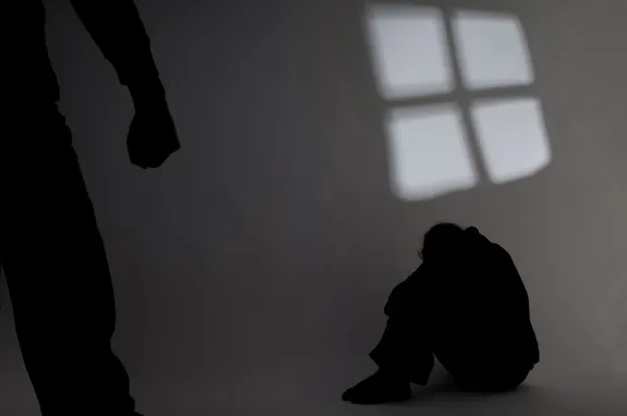 Two shadowy figures. One is curled in a ball on the floor, protecting themselves with their hands. The other figure is stood, we can see their arm and clenched fist.