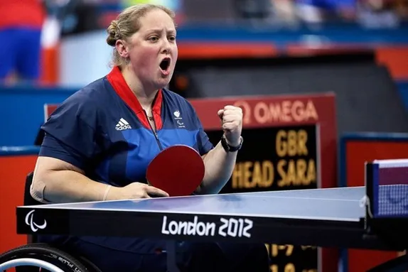 Sara is sat a table tennis court in her wheelchair. She holding a table tennis bat in one hand and is punching the air in celebration with her other hand. The table has the ‘London 2012’ logo on it.