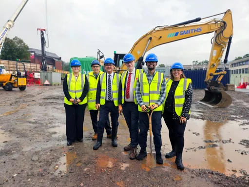 Mae grŵp o chwech o bobl, sy'n gwisgo hetiau caled glas a siacedi melyn hi-vis, yn sefyll ar ddarn o dir mwdlyd o flaen cloddiwr mawr. Mae un o'r bobl yn dal sbri.