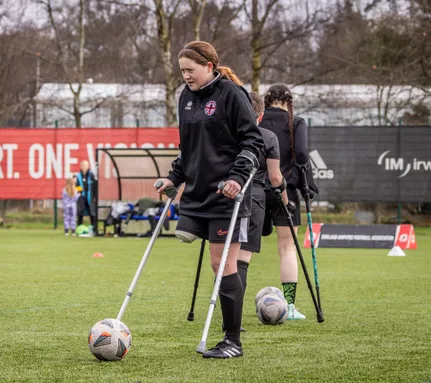 Image of Tayla on a pitch with a football on the ground. She is wearing a black football kit and using crutches.