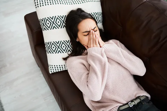 The image shows a woman lying on a couch, resting her head on a decorative pillow with geometric patterns. She is wearing a light pink sweater and has her hands covering her face, as if she is upset, stressed, or tired. The overall mood suggests she may be experiencing emotional distress or exhaustion. The couch appears to be dark brown, and the floor is light-coloured, creating a contrast in the