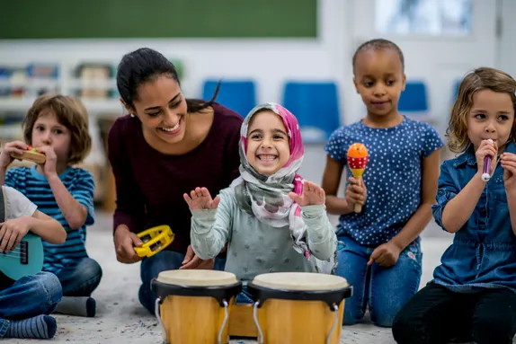 A group of young children and an adult are joyfully playing musical instruments together in a classroom. The children are using instruments such as drums, maracas, a small flute, and a ukulele. The adult, sitting in the centre, is smiling and holding a tambourine, engaging with the children. One child wearing a colourful hijab is happily playing the drums, smiling brightly. The setting is bright a