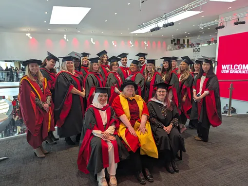 A group of graduates and faculty members posing for a photo indoors at a graduation ceremony.