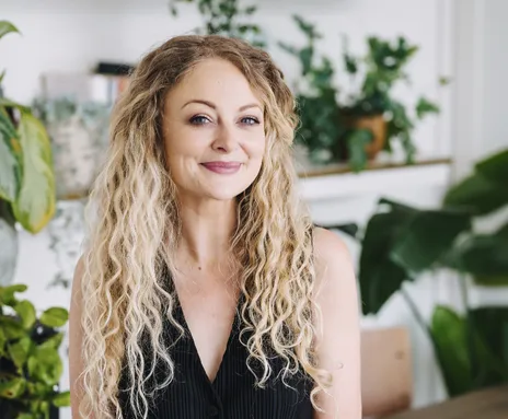 Kayley is smiling. She has long, curly blonde hair and wearing a black sleeveless top, sitting in a room filled with lush green plants and light-coloured decor.