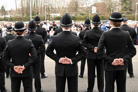 A group of police officers in formal black uniforms and traditional helmets stand in formation with their hands clasped behind their backs. They are facing a crowd of spectators, who are gathered outdoors in a public space. The setting includes trees, lampposts, and parked cars in the background, indicating an official passing-out parade.