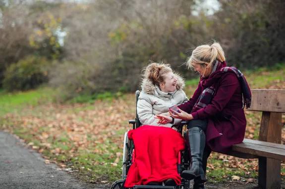 Two people sitting outdoors in a park on a fall day. One woman, wearing a white coat, is in a wheelchair covered with a red blanket, smiling and looking at the other woman sitting beside her on a bench. The second woman, wearing a burgundy coat and scarf, is holding a mobile phone and engaging warmly with her companion. The background features a pathway, bare trees, and fallen leaves.