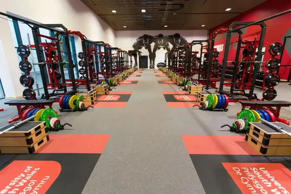 A modern, well-equipped strength and conditioning gym at the University of South Wales, featuring rows of red squat racks, Olympic barbells with colourful weight plates, wooden plyometric boxes, and a mural of sprinters on the far wall.