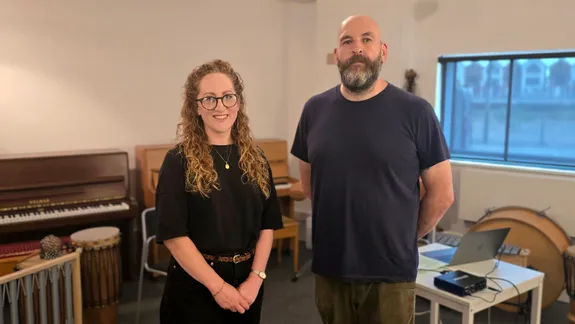 Mae menyw gyda gwallt hir, cyrliog, sbectol, a gwisg ddu yn sefyll yn gwenu wrth ochr dyn â phen heb wallt, barf lawn, a siwt t tywyll. Maent mewn ystafell gerddoriaeth gyda chynnau offer cerddorol yn weladwy, gan gynnwys piano, drymiau, a gliniadur ar fwrdd yn y cefndir.