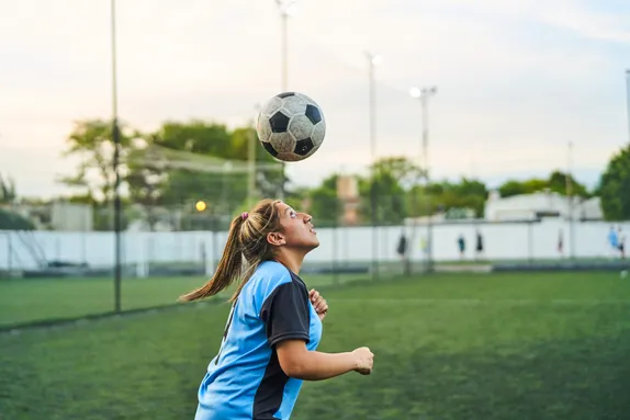 A female footballer, wearing a blue and black top, heads a football. They are standing on a football pitch.