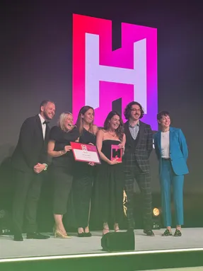 A group of six people dressed in formal attire smile on stage at an awards ceremony. Two women in the centre hold a certificate and a pink trophy shaped like the letter H. Behind them is a large illuminated logo featuring a bold white ‘H’ on a pink and purple background.
