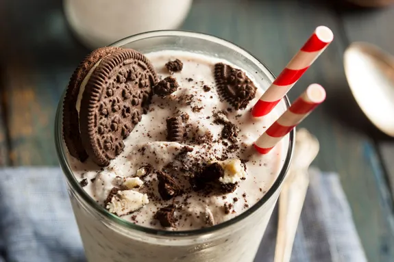 A cookies and cream milkshake sits on a wooden table in a tall glass with a spoon beside it. There is a large biscuit in the milkshake along with crumbled chocolate cookies. There are two red-and-white straws standing in the glass.