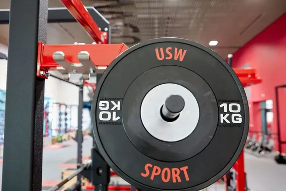 A close-up of a 10kg black weight plate labelled USW Sport on a red and black weightlifting rack.