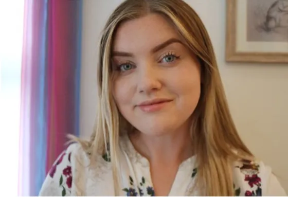 A young woman with long blonde hair smiles softly at the camera. She is wearing a white floral top, and behind her is a softly coloured curtain and a framed picture on the wall.