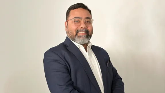 A person wearing a dark blazer over a white shirt, standing against a plain light-coloured background.