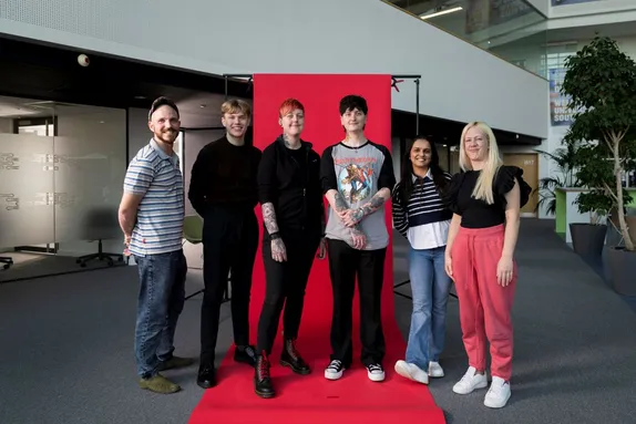 Six people stand side by side on a red backdrop in a modern indoor space, smiling at the camera.