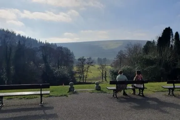 Safle awyr agored hardd gyda sawl banc pren wedi'u trefnu ar hyd llwybr paentiedig. Mae ambell i berson yn eistedd ar y banciau, gan edrych ymlaen at dirwedd eang o gaeau gwyrdd, coed wedi'u gwasgaru, a bryniau pell.