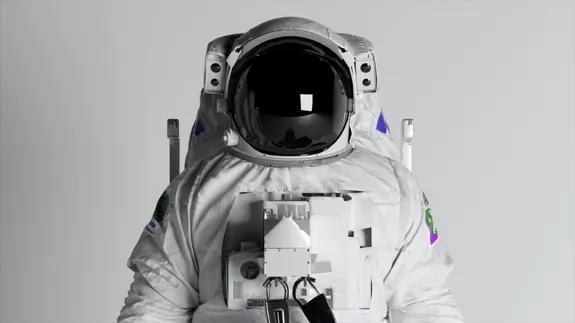 Front-facing view of an astronaut in a white spacesuit against a plain light-grey background. The helmet visor is black and reflective.