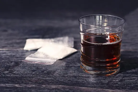 A glass of dark liquid on a wooden surface beside two small clear plastic bags containing white powder.