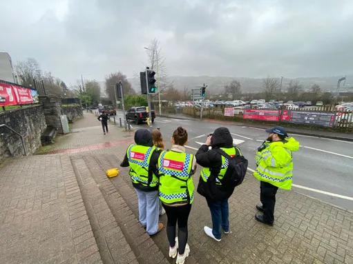 Four students wearing high-visibility vests stand on a pavement beside a police community support officer in a fluorescent jacket, observing a roadside speed monitoring operation at a traffic light junction. One person appears to be using a handheld speed detection device while cars pass.