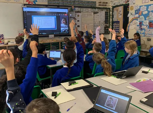 School pupils raise their hands to a teacher on a digital screen during a class at NDEC
