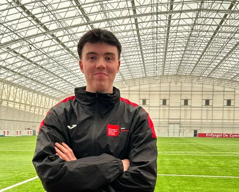 A football coaching student wearing a University of South Wales Sport jacket stands with arms crossed on an indoor artificial pitch, inside a large covered training facility.